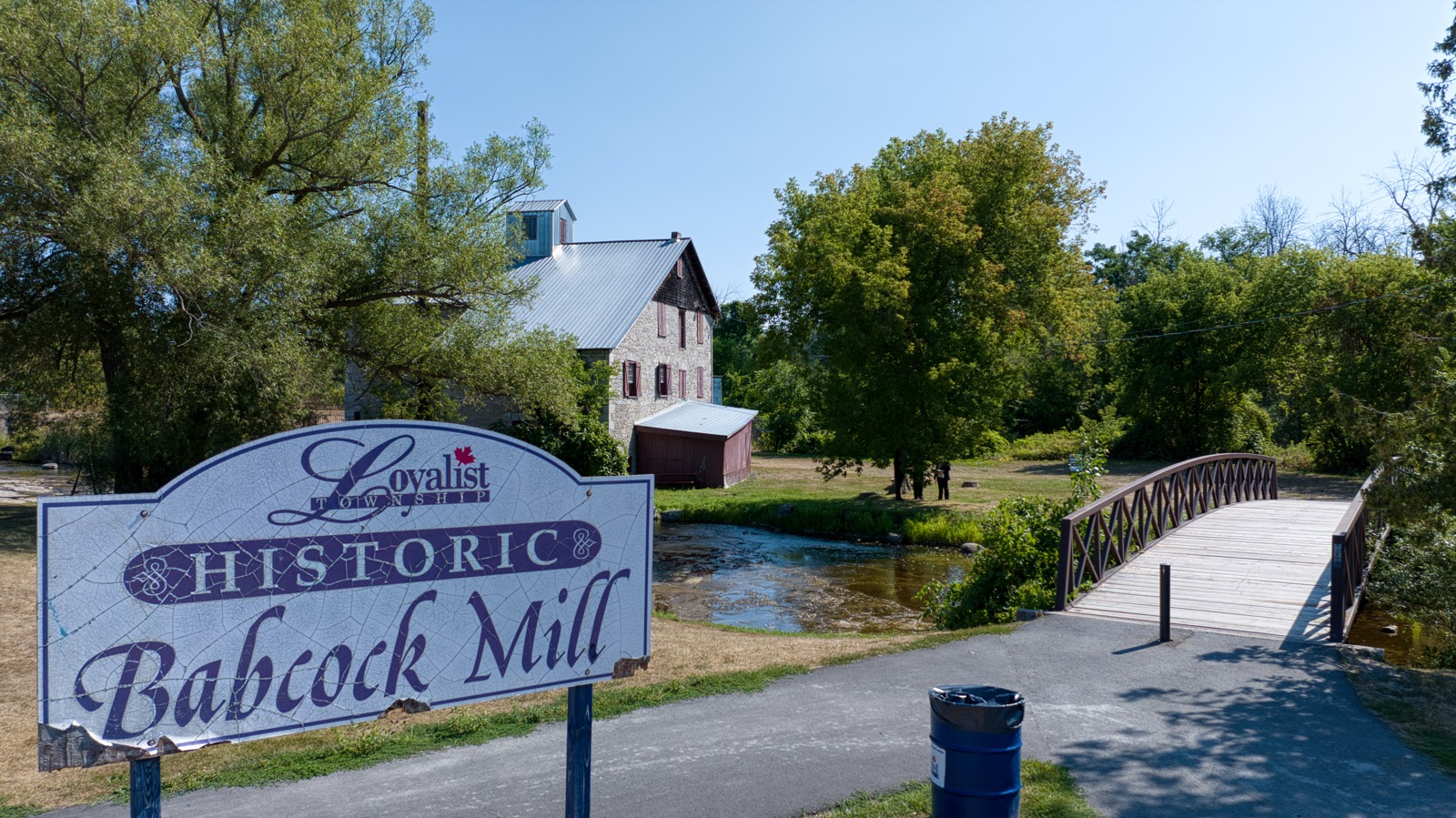The stone mill and wooden footbridge at Babcock Mill, with the Loyalist Township sign in front.