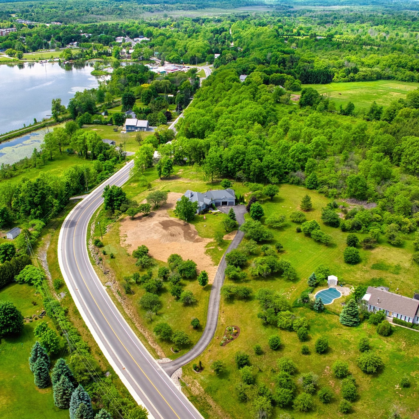 Aerial of a house, a cleared yard, and a curving road beside woods and water.