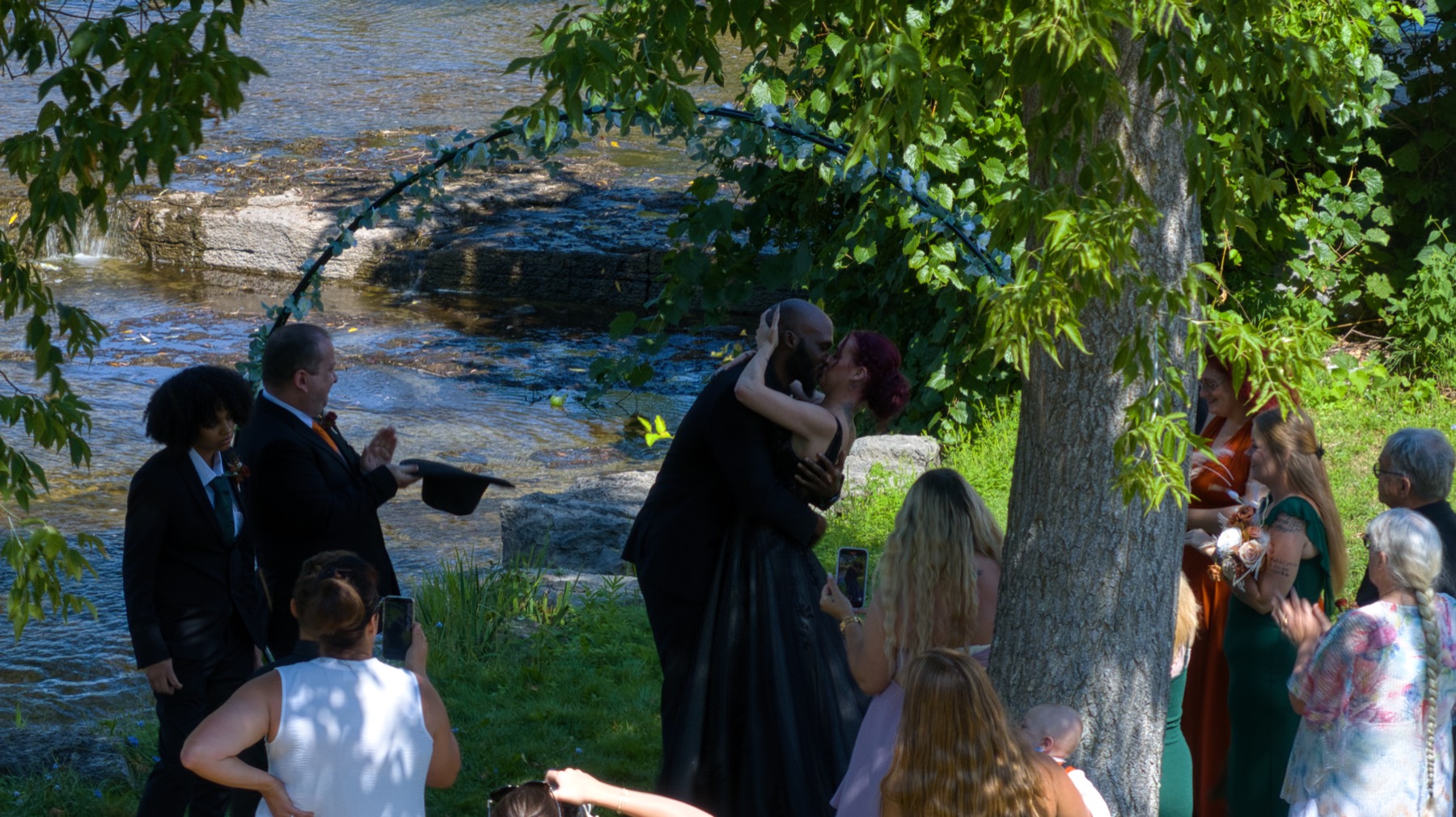 The couple kissing under a floral arch beside the creek, guests clapping.